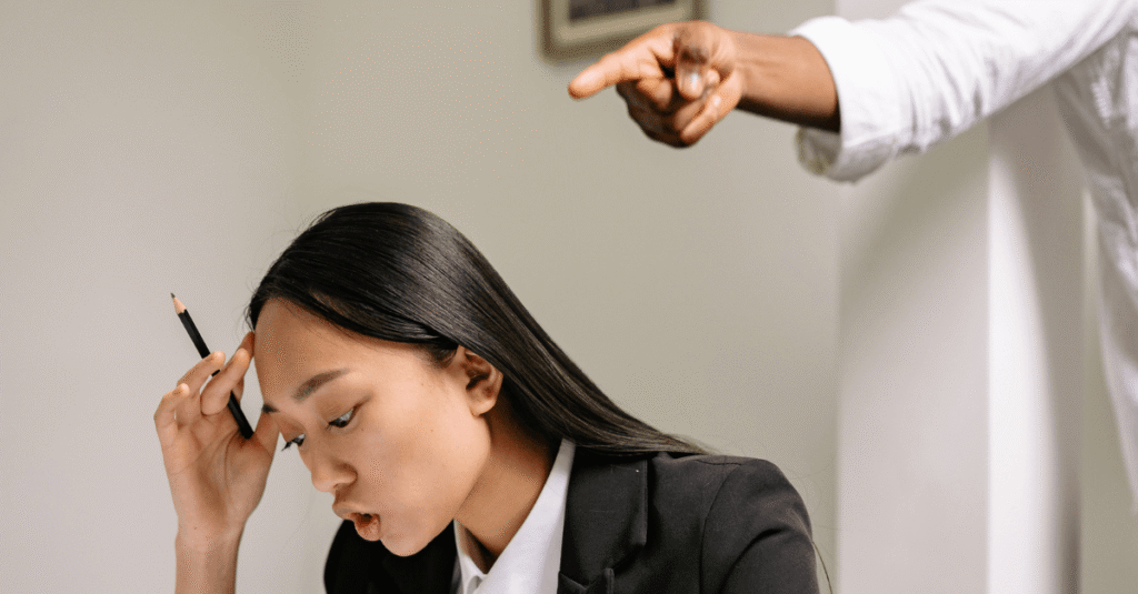 A women sitting and looking at a paper with a pen in her right hand near hear head. Someone behind her is pointing at her.